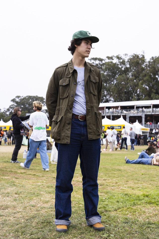 A young man wearing a green cap, olive jacket, gray shirt, dark jeans, and brown shoes stands on grass at an outdoor event with people and tents in the background.